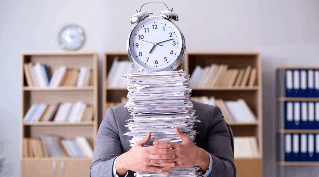 Person's head replaced by an alarm clock, buried under a towering stack of papers, in an office setting.