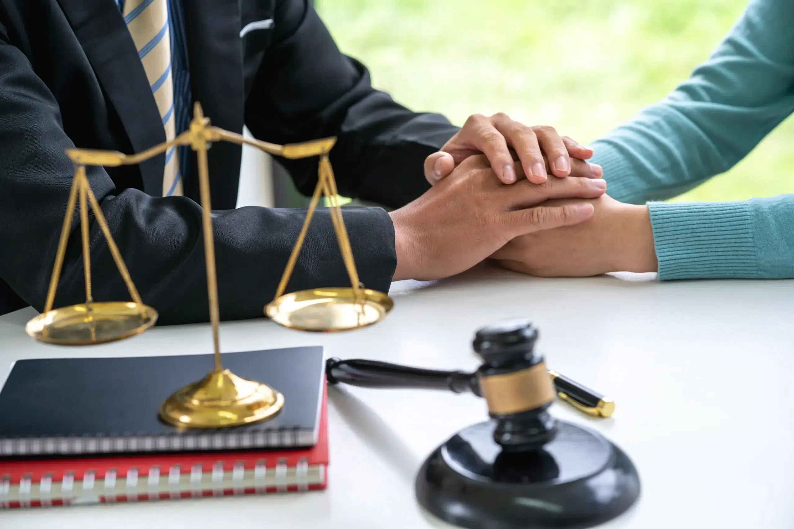 Lawyer's hands comforting a client's hands on a table with scales of justice and a gavel, symbolizing legal support.
